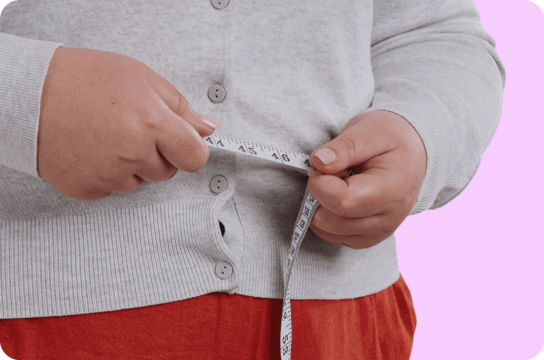 Person measuring their waist with a tape measure over a grey sweater, standing against a pink background.