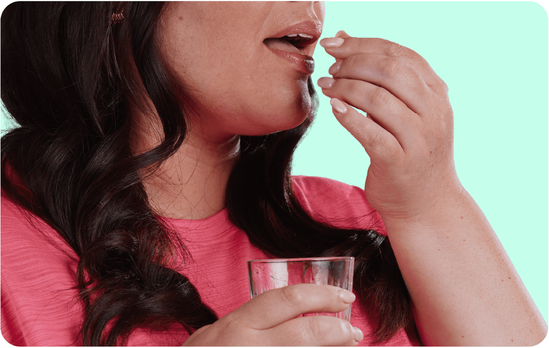 Woman in pink shirt taking a pill with a glass of water, set against a light green background.