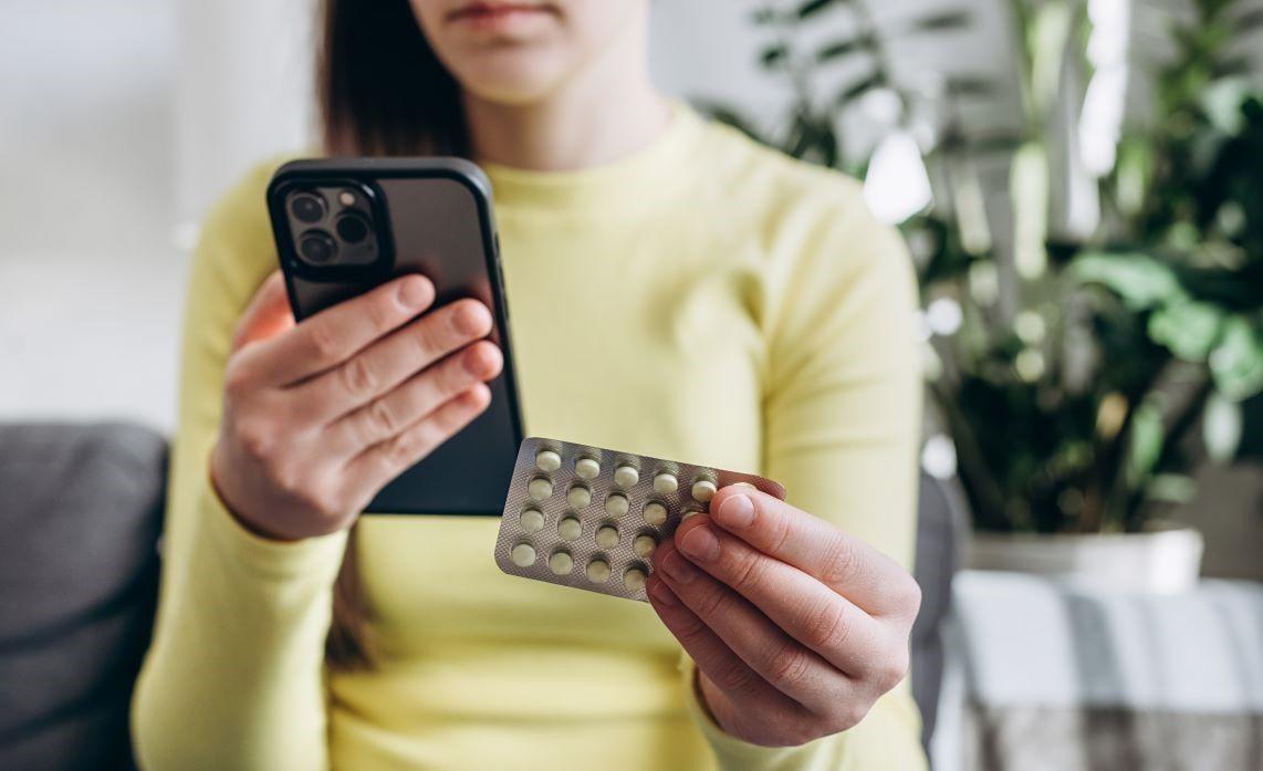 A young woman in a yellow jumper holding a blister pack of medicine and checking information about it on her phone