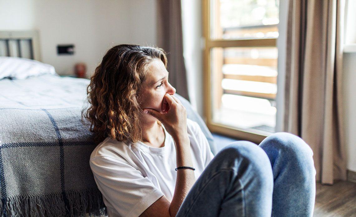Woman sitting on the floor next to a bed, looking thoughtfully out a window, with sunlight streaming in through sheer curtains