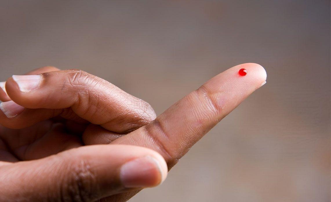 A close-up of a hand with a drop of blood on the tip of the index finger, set against a neutral background