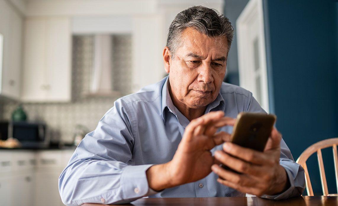 Man in a light blue shirt sitting at a table in a kitchen, focused on using a smartphone with both hands