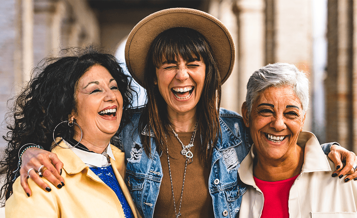 Gen M and MTick Menopause help at Pharmacy2U. Three women standing close, laughing joyfully. The center woman wears a hat and denim jacket, while the others are in light jackets. Indoor setting.