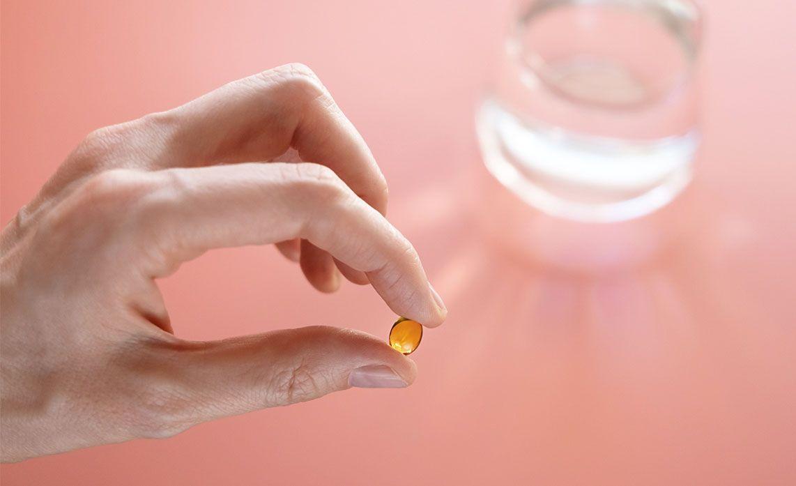 Hand holding a small orange pill between fingers, with a glass of water in the background on a pink surface.