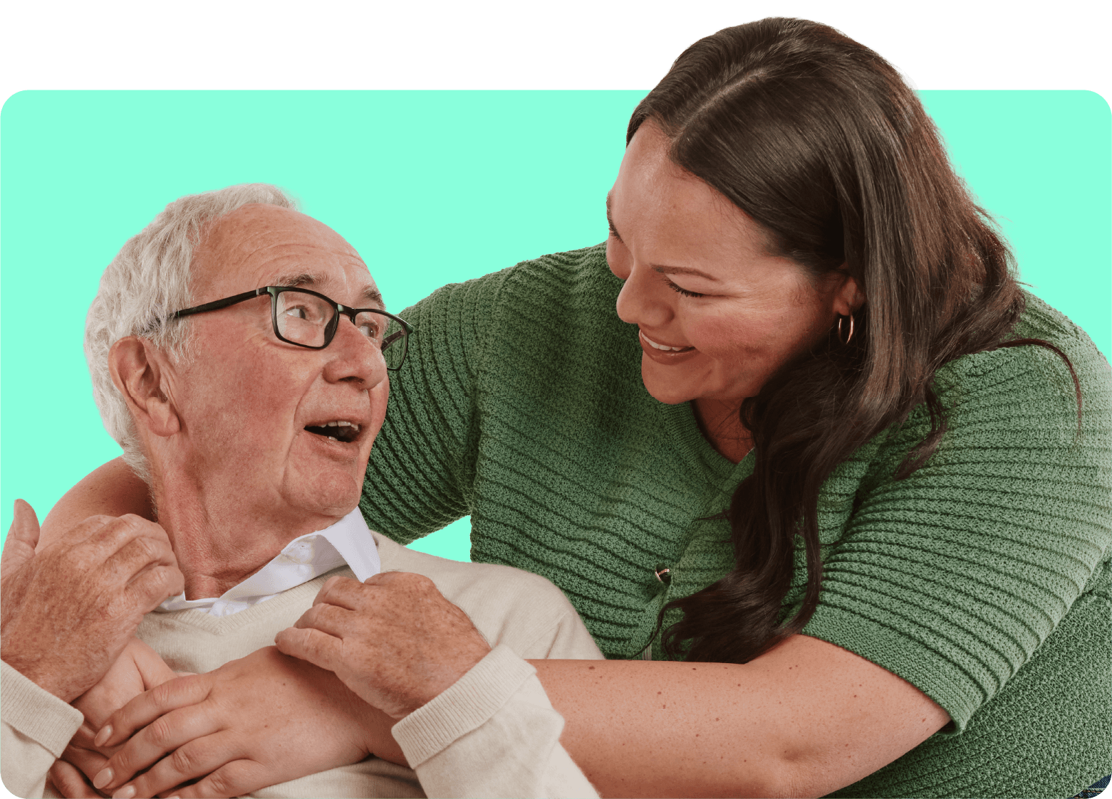 Elderly man smiling at a woman who hugs him warmly from behind against a teal background.