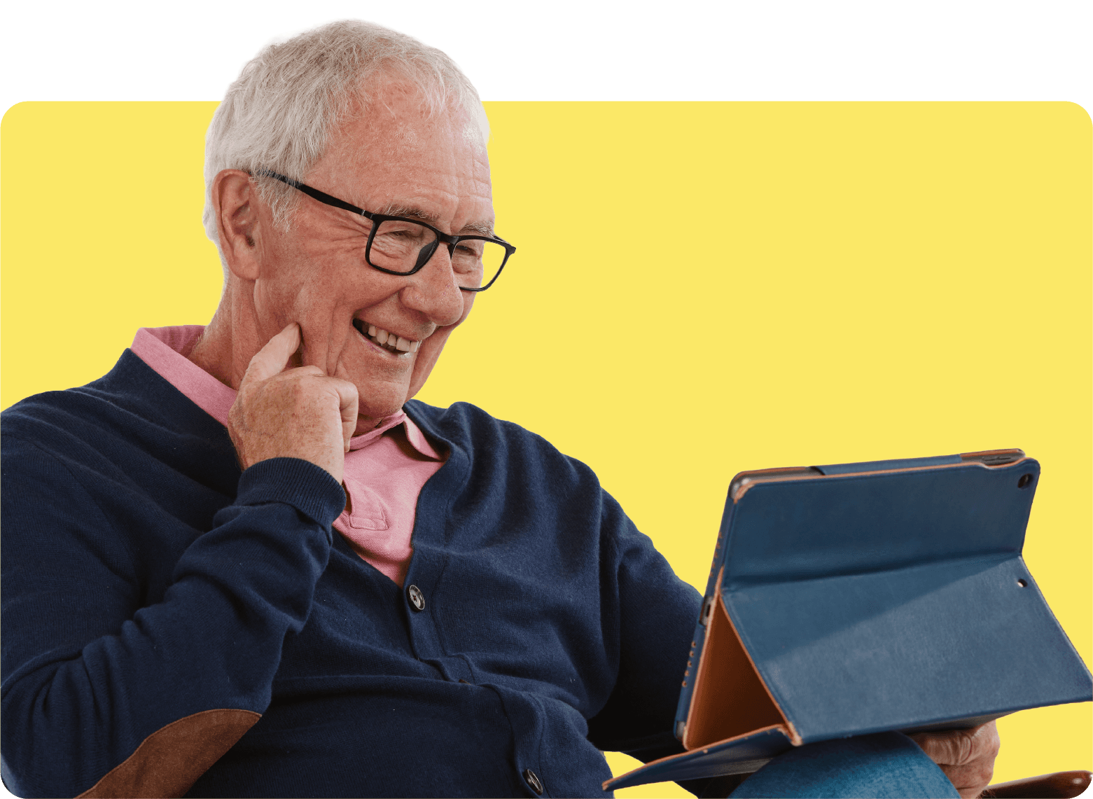 Elderly man with glasses smiles while using a tablet, wearing a navy cardigan and pink shirt, against a yellow background.