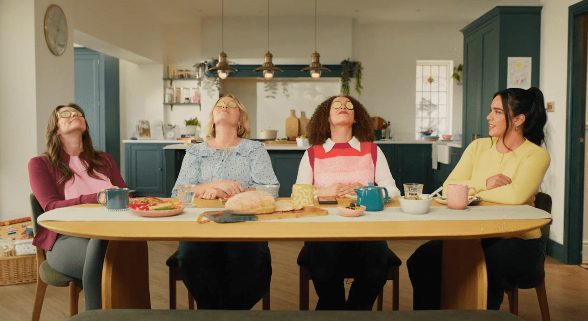 Four women sit at a table, three with cucumber slices on their eyes, enjoying a relaxing moment in a cosy kitchen with snacks and drinks