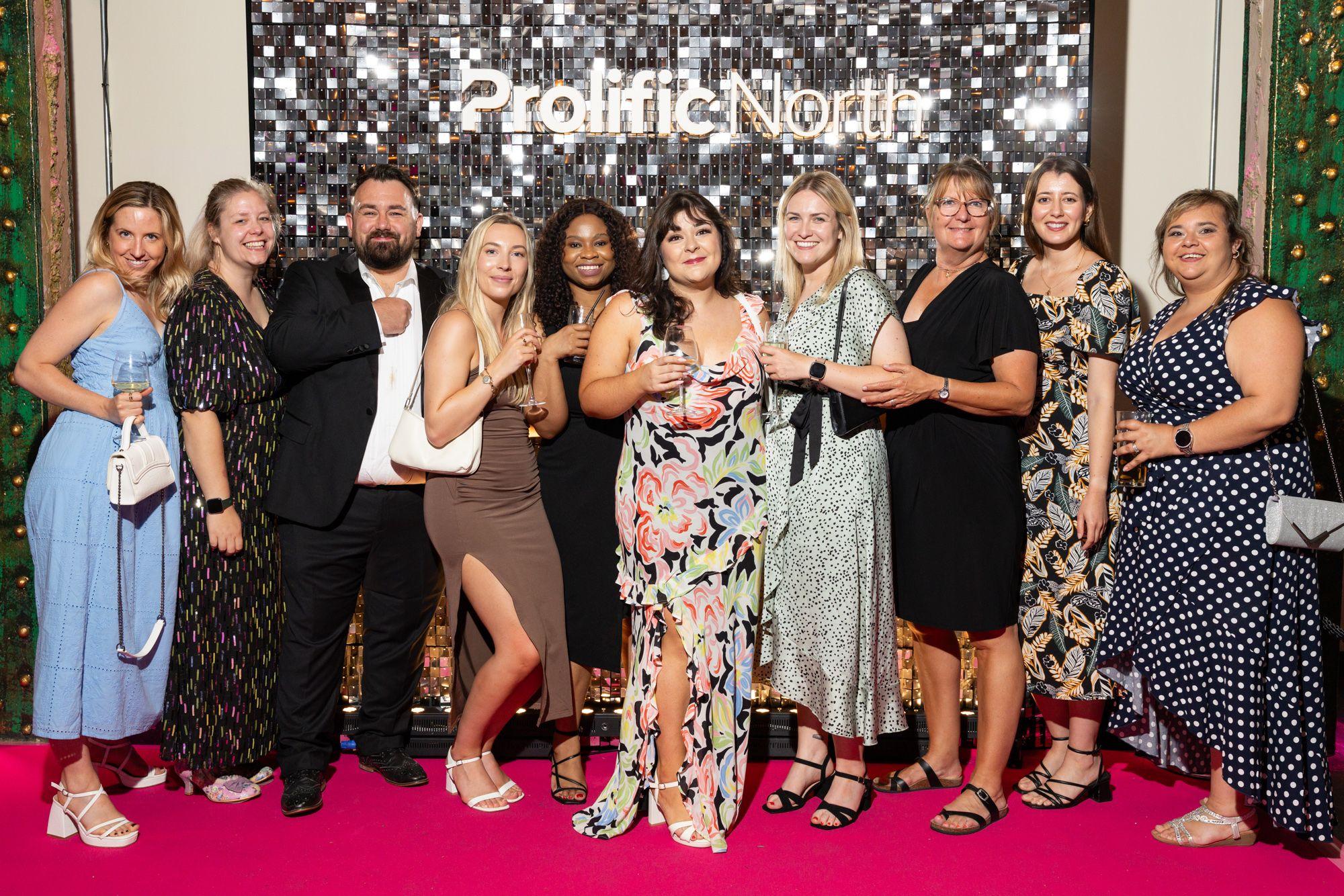 A group of people dressed in semi-formal attire pose together on a pink carpet in front of a "Prolific North" backdrop.