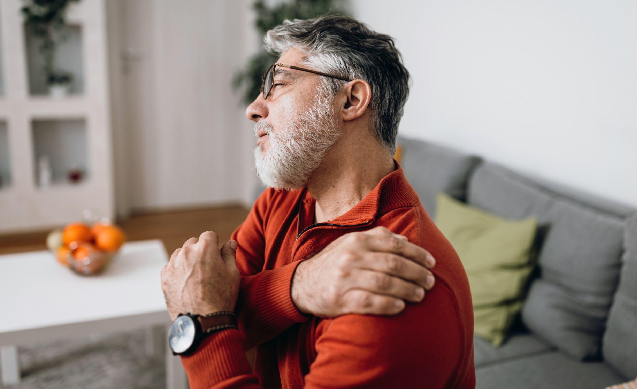 Older man in a red sweater sits on a couch, holding his shoulder and appearing to experience pain. A table with a bowl of oranges is in the background.
