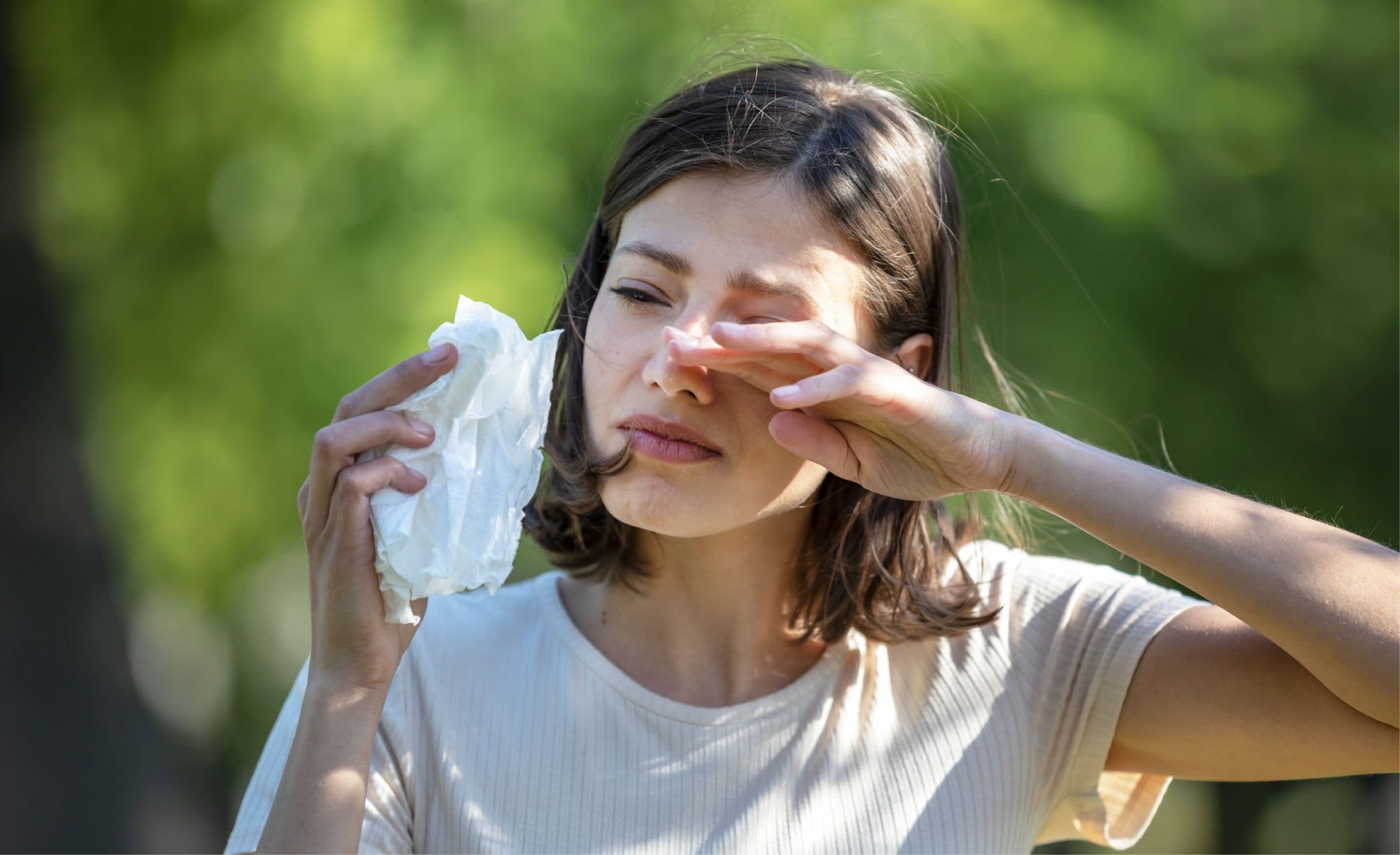 Woman wiping her teary eyes with a tissue, appearing to have allergies, standing outdoors.