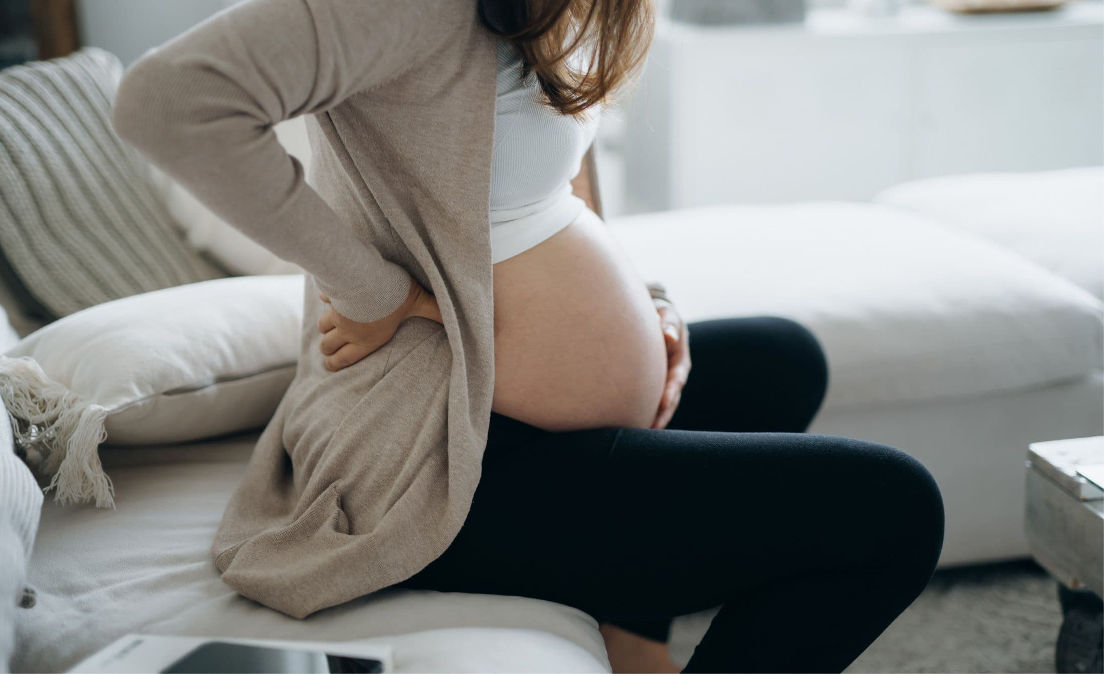 Pregnant woman sitting on a couch, holding her lower back, wearing a light cardigan and black pants in a cozy living room setting.