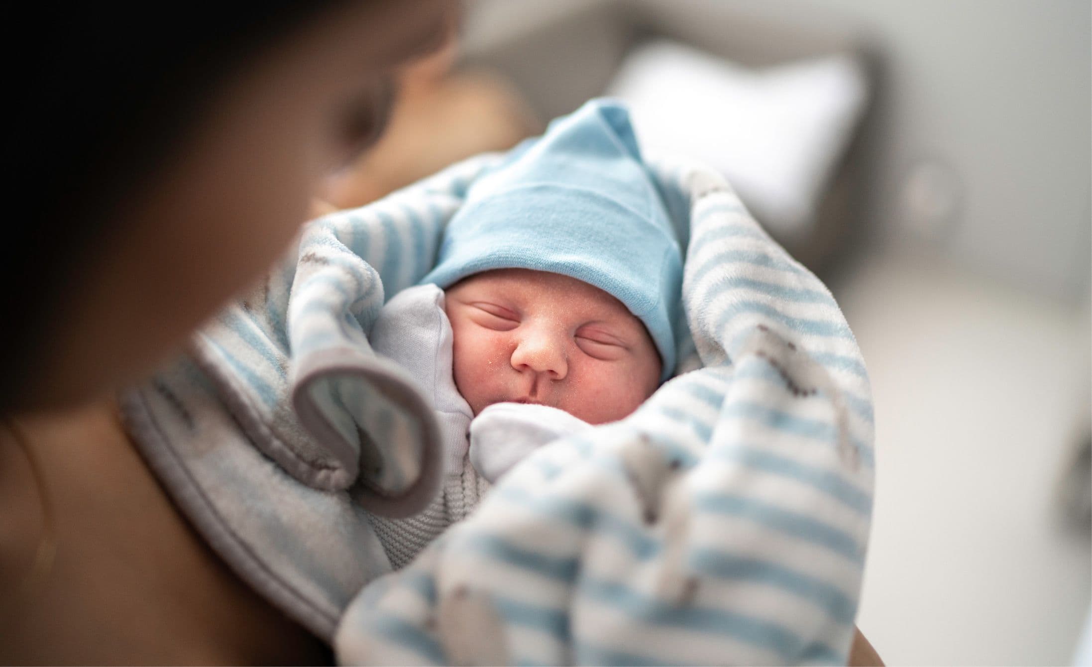 A newborn baby wrapped in a blue and white striped blanket wears a blue hat, peacefully sleeping in an adult’s arms.