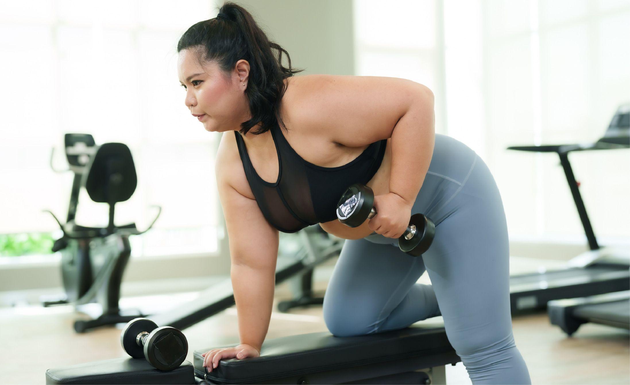Woman lifting a dumbbell in a gym, leaning on a bench with focus, wearing a black top and blue leggings. Exercise equipment is visible in the background.