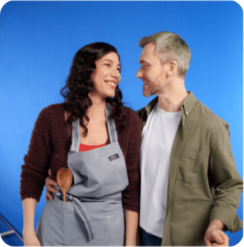 A woman in an apron smiles at a man in a green shirt against a blue background. They appear happy and affectionate.