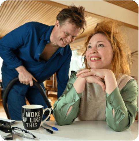 A woman with messy hair smiles at a table, while a man with tousled hair vacuums behind her. A mug reads, "I woke up like this."