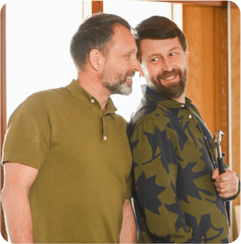 Two men smiling at each other indoors; one holds a hammer, wearing a green patterned shirt, the other in a plain green polo shirt.